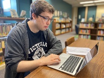 A student wearing a Victoria College shirt and glasses sits in the library with a laptop
