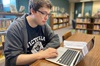 A student wearing a Victoria College shirt and glasses sits in the library with a laptop