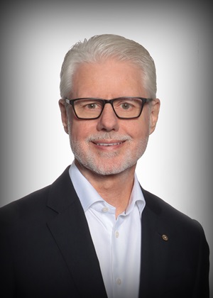 Phil Green smiles wearing glasses and a black blazer in front of a white backdrop