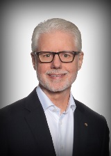 Phil Green smiles wearing glasses and a black blazer in front of a white backdrop