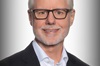 Phil Green smiles wearing glasses and a black blazer in front of a white backdrop